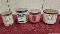 Four scented candles arranged side by side on red fabric. Various colors and metal lids, different brands visible.