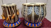 Overview of both tabla drums (wooden dayan left, metal bayan right) on silk cushions with tuning hammer leaning against dayan