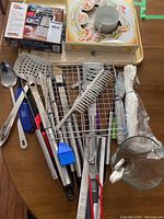 Various BBQ and kitchen tools including metal spatula, brush, tongs, cleaning comb, silicone brush, metal fork, trays and glass measuring cup.