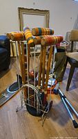Full view of the croquet mallets with striped wooden heads and handles placed in a metal stand on a wooden floor.