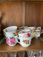 Image showing a collection of 6 floral porcelain cups on a wooden shelf with various rose patterns, mostly pink and yellow with gold trims.