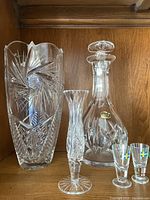 Photo showing decanter with stopper, tall crystal vase, and two Swedish flag shot glasses on wooden shelf