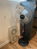 Photo showing both fans: Uberhaus pedestal fan and Hampton Bay dual window fan leaning against wall.