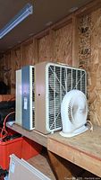 Three box fans and one white Airworks desk fan on wooden shelves in a workshop setting.