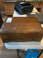 Closed wooden flatware chest with a curved top and bottom drawer, placed on a table.