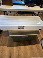 Two storage bins stacked on floor, including a transparent white under-bed storage container and a green storage bin.