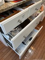 Photo of three open drawers displaying an assortment of kitchen cooking utensils and pots/pans inside a white cabinet under a countertop.