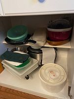 Several kitchen items grouped on and under a shelf including bowls, frying pan, plastic lids, paper towel roll, and a metallic crock pot.
