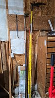 Extended branch trimmer and long pole tools leaning against a wall in a wood shed or storage