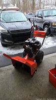 Front angle of snow blower showing red two-stage auger housing, wheels, engine and extension cord on handle
