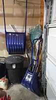 Wide view showing two large blue plastic snow shovels, black garbage can, green garden rake, and several cleaning tools with wooden handles hung along the wall.