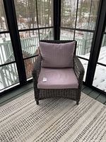 Front view of one dark woven wicker arm chair with light gray cushions in porch setting with snowy background.