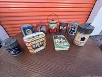 Photo of 10 assorted vintage collectible tins in various shapes and designs, placed on ground in front of orange storage unit door.