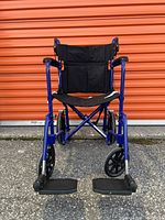 Front view of Excel transport wheelchair showing black nylon seat and backrest, blue aluminum frame, and detachable footrests with textured foot platforms.