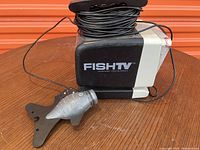 Photo showing Fish TV branded control unit with attached long black cable and silver fish-shaped weight/mount on wooden table surface.