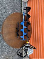 Top view of the Jiffy 2500 ice fishing auger motor unit attached to a long black auger bit, lying on a circular wooden surface outdoors with two additional drill bits - one blue medium-sized and one smaller black bit.