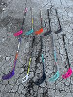 Photo of 10 Salming floorball sticks laid out on asphalt, showing a variety of blade colors including pink, turquoise, orange, purple, and black with shafts of different colors and graphics.
