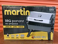 Photo of original box showing branding and key features like 14,000 BTU burner, folding legs, tubular burner, and porcelain cooking grid.