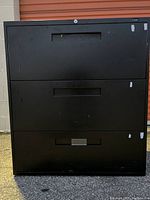 Front view of a black three drawer solid steel file cabinet with some visible scratches, lock cylinder present but no key.