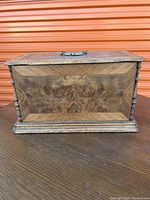 Front view of closed ornately veneered wooden case with metal handle showing wood grain and carved details.