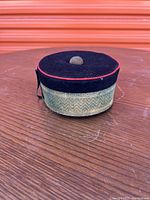 Front and top view of black pill box hat with red piping, gold woven band, and fabric button on top.
