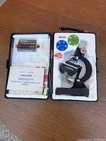 Open carrying case showing the microscope, prepared slides, lens paper, and colored filters placed in foam compartments.