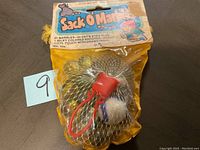 Unopened vintage 1980 Sack O'Marbles with marbles visible inside yellow mesh pouch showing orange drawstring and plastic stopper, attached to branded colorful header card.