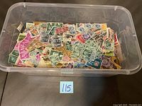 Top view of a transparent plastic container showing a large collection of assorted used stamps inside.