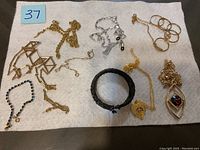 Overview of all jewelry items laid on a white cloth showing multiple gold-tone chains, silver-tone chain, rings, black braided bracelet, and beaded blue bracelet.