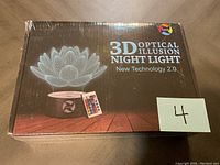 Photo of the sealed box showing the 3D Optical Illusion Night Light featuring a lotus flower design and a remote control. The box has the brand Luxe and states New Technology 2.0.