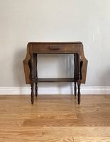 Full front view of the antique wooden side table showing drawer, drop-leaf sides, and turned legs.