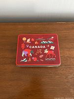 Red metal box with colorful Canadian themed graphics on the lid, closed, showing the top design.
