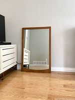 Front view of mid-century walnut framed mirror leaning against wall, showing overall condition and size relative to floor and nearby furniture.