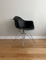 Side angled view of black modern plastic armchair with chrome Eiffel-style metal legs on wooden floor against plain wall.