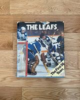 Front cover of 'The Leafs The First 50 Years' book showing a hockey game with players in blue and white, some in the net and others skating, with a title and visible rips on the dust jacket.