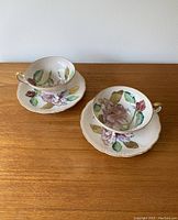 Photo showing two fine porcelain teacups and matching saucers with hand painted pink flowers, green leaves, and gold trim on handles and rims, displayed on wooden surface.
