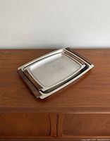 Two stacked trays made of stainless steel with teak wood handles shown on wooden cabinet. Smaller tray nested inside larger one.