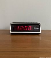 Front view of the Dynasty Dream Machine clock displaying 12:00 with red LED numbers, white frame, and black face with Dynasty branding.