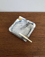 Angle view of the square glass ashtray showing marbled black and white pattern with a cigarette resting in one of the corner indentations with gold accents.