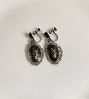 Photo shows front view of the vintage clip-on Siam sterling silver earrings with black oval and silver figure inlays, decorative silver edges, and clip screw backs.