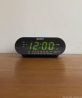 Front view showing green LED display active with 12:00 time, Sony logo, AM/FM frequency scale and alarm indicator.