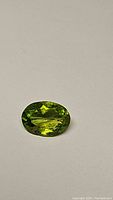 Close-up photo showing the apple green oval cut peridot gemstone on a plain background, highlighting the cut, color and clarity.