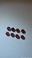 Eight small natural round cabochon sapphires in raspberry pink to red shades, laid out on a flat light background, showing their smooth domed surfaces.