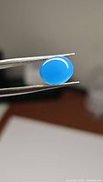 Close-up image of natural oval cabochon lagoon blue chalcedony gemstone held in tweezers showing bright lagoon blue color and smooth polished surface.