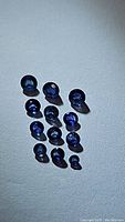 Thirteen natural royal blue sapphires arranged on a light surface, showing their rich blue color and round faceted cuts under lighting.