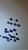 Photo shows multiple small round cut blue sapphire gemstones scattered on a white surface, varying in size.