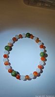 Top down photo of a round stretchy bracelet composed of polished beads in white, green, and orange-red colors placed on plain surface.