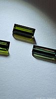 Close-up photo of three baguette-cut green tourmaline gemstones on white surface showing rectangular facets and clarity.