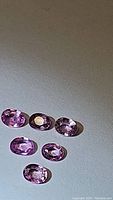 Photo of six oval cut natural pink sapphires arranged on flat surface under light showing pink hues and luster.