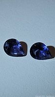 Two pear-shaped deep purple iolite gemstones on gray background, showing facets and color under bright light.